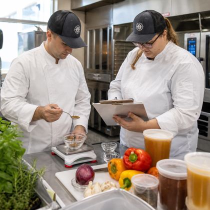 Two chefs weighing and tasting a sauce alongside fresh peppers, herbs and stock in the test kitchen