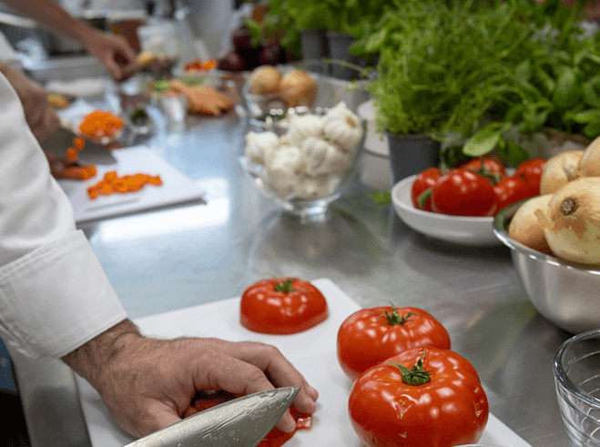 A cook slicing tomatoes on a cutting board beside bowls of garlic, onions and fresh herbs
