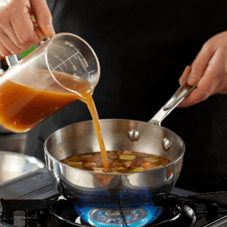 Broth concentrate being poured from a measuring cup into a saucepan over an open flame