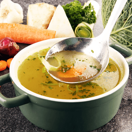 A ladle lifting vegetable broth from a green pot surrounded by carrots, leeks and cabbage
