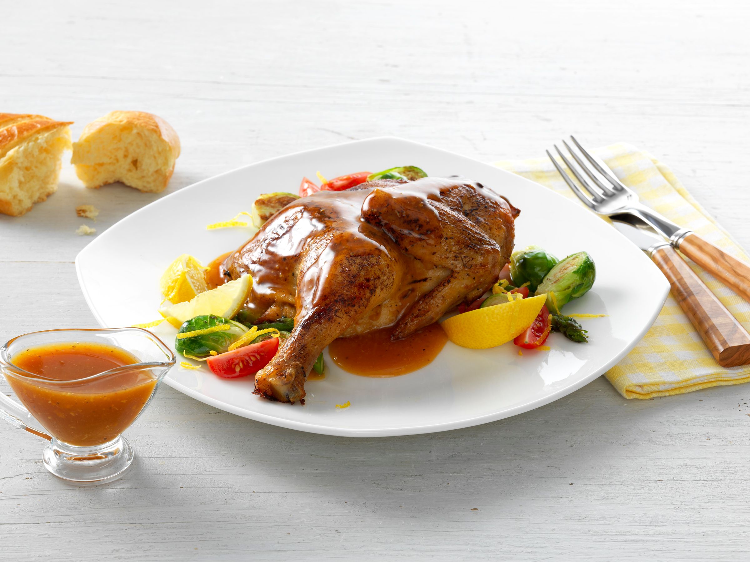 A roast chicken entrée plated with brussels sprouts, tomato and lemon, with a boat of pan sauce alongside