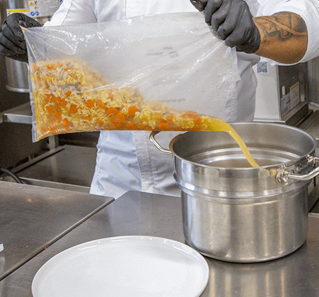 A cook emptying a ready-to-use pouch of chicken noodle soup into a stockpot