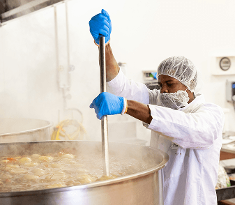 A production cook stirring a steaming kettle of soup with a long paddle