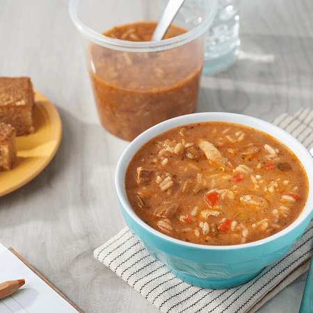 A bowl of chicken and sausage gumbo beside a ready-to-eat cup and a plate of cornbread